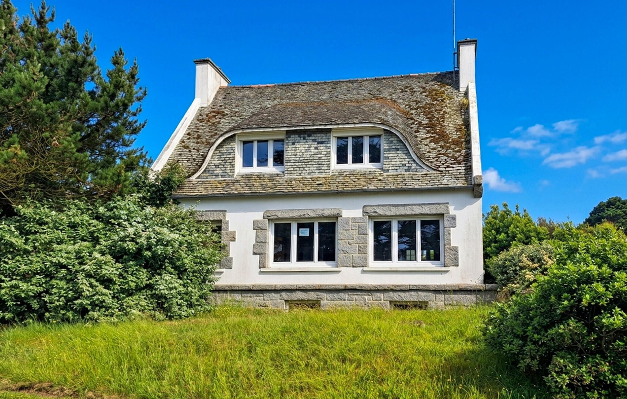 Une maison à deux étages, avec un toit en pierre, des murs blancs et de nombreuses fenêtres.