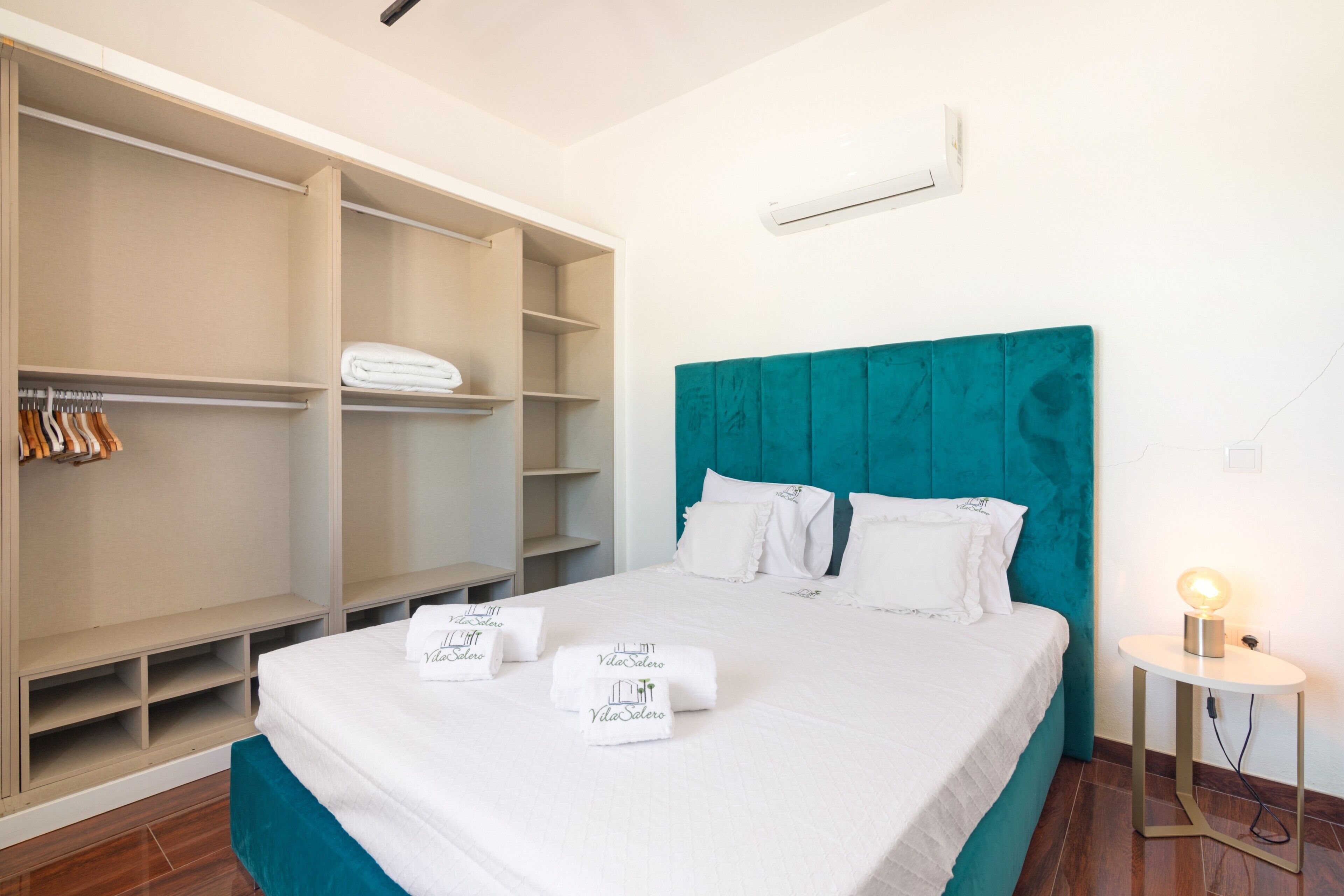 A hotel room with a bed, a teal headboard, a wardrobe, and a bedside table with a lamp.