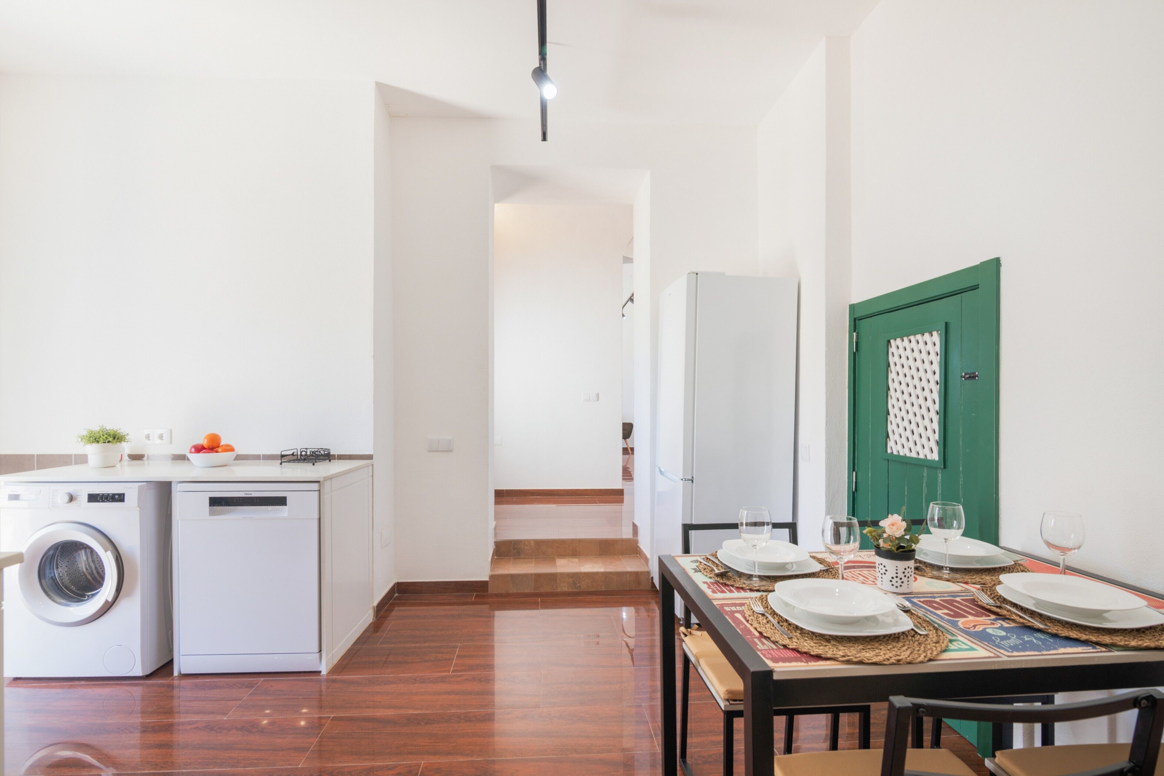 A compact kitchen with a washing machine, a dining area with a table set for two, and a green door.