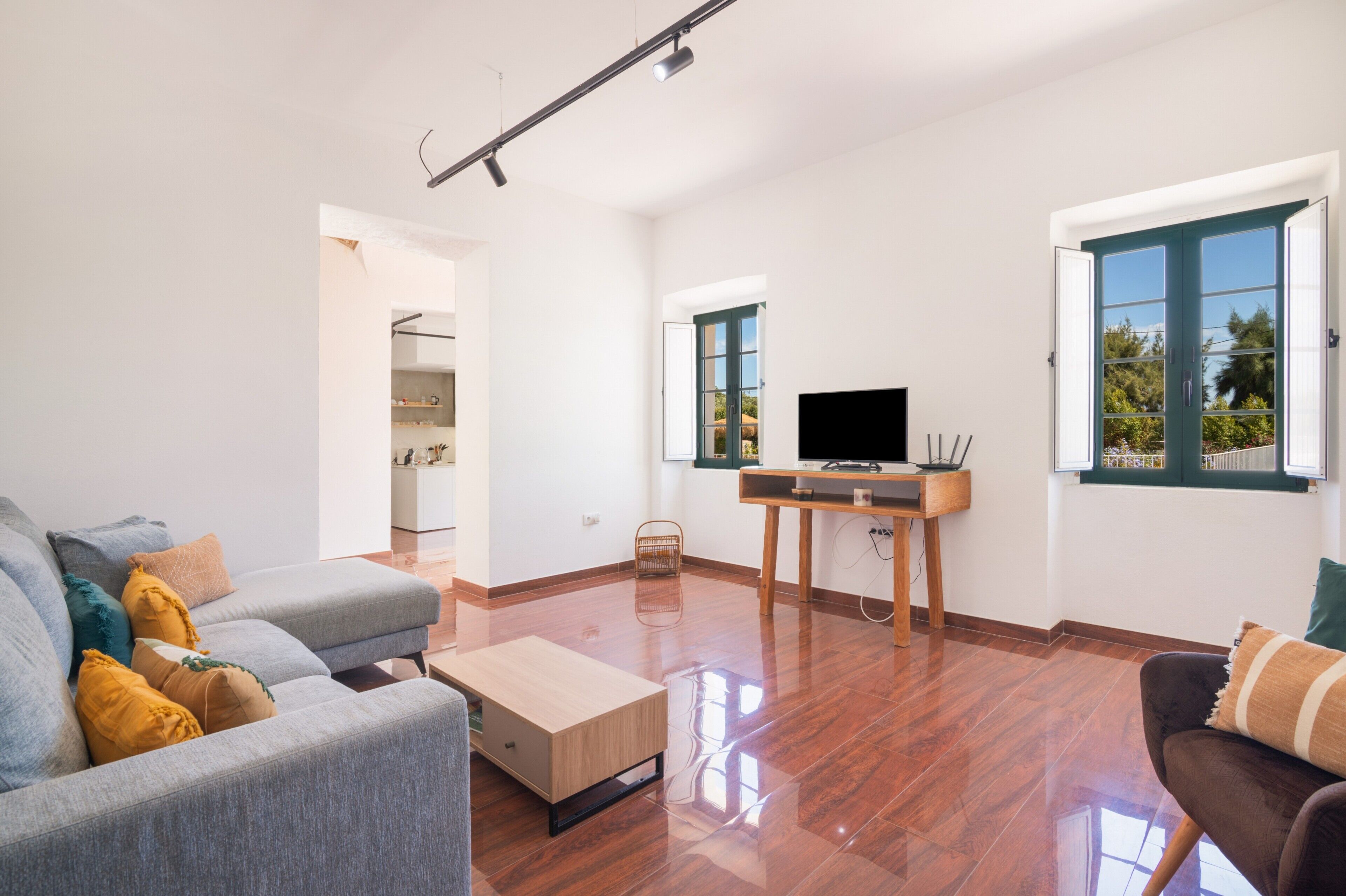 A spacious living room with a grey sofa, a wooden coffee table, a TV on a stand, and a window with a view of greenery.