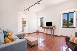 Ett rymligt vardagsrum med en grå soffa, ett träbord, en tv på ett tv-bord och ett fönster med utsikt över grönområden.