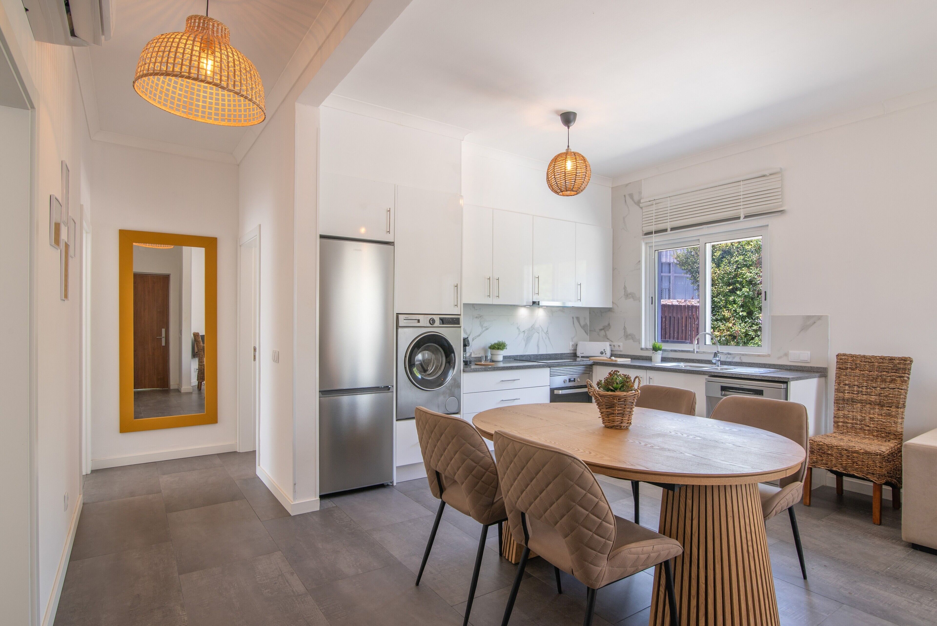 A modern kitchen with a stainless steel refrigerator, a washing machine, and a dining area with a wooden table and cushioned chairs.