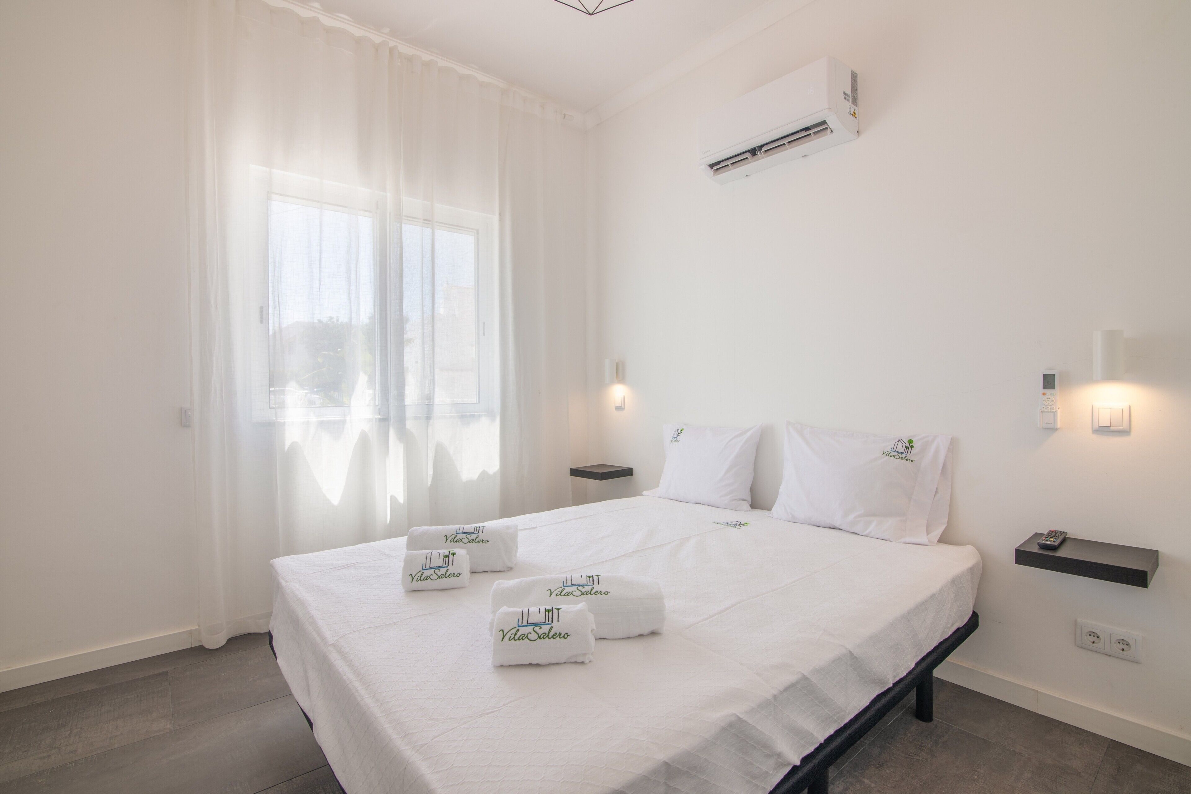 Hotelkamer met een bed, nachtkastje, airconditioning en een raam met dunne gordijnen.