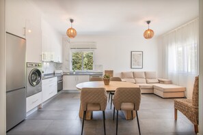 Uma cozinha moderna com máquina de lavar embutida, sala de jantar com mesa redonda e cadeiras acolchoadas, e sala de estar com sofá e poltrona de vime.