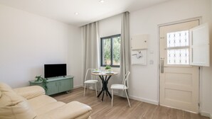 Ein modernes Wohnzimmer mit einer Couch, einem Fernseher auf einem Schrank, einem Esstisch mit Stühlen, einem Fenster mit Vorhängen und einer Tür mit Fenster.