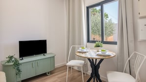 Ein Zimmer mit Fernseher auf einem grünen Schrank, einem Esstisch mit Stühlen und einem Fenster mit Vorhängen.