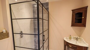 Ein Badezimmer mit Duschkabine aus Glas, Marmorboden, Waschtischunterschrank aus Holz und einem Holzschrank über dem Spiegel.