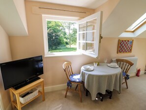 A room with a TV on a wooden stand, a dining table with chairs, a window with an open shutter, and a radiator. - The Stable Flat (Lutterworth)