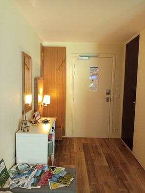 A hotel room with a wooden floor, a desk with a lamp, a mirror, and a door with a bulletin board. - The Little Refuge (Les Déserts)