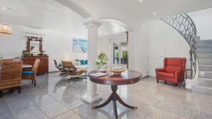 Um hall amplo com uma mesa redonda, uma poltrona de couro vermelha, uma escadaria e um espaço de refeições com móveis de vime.