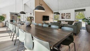 Un vaste espace dédié à la salle à manger, avec une longue table en bois, des chaises modernes, un éclairage suspendu, un téléviseur et une grande fenêtre donnant sur un paysage verdoyant.