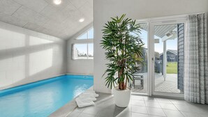 Une piscine intérieure agrémentée d’une grande plante en pot, une porte-fenêtre coulissante donnant sur l’extérieur et un sol carrelé.