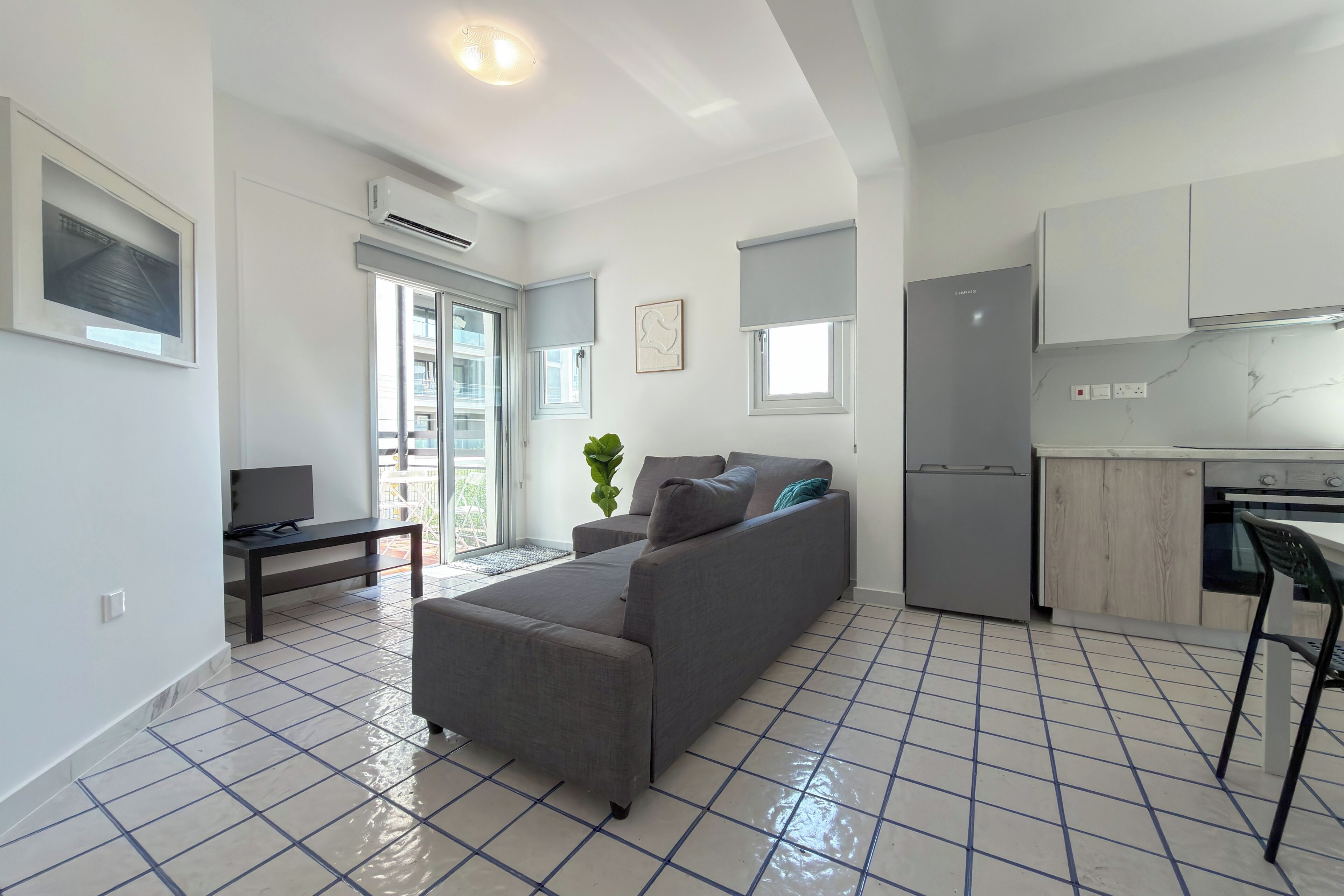 Una sala de estar moderna con un sofá gris, una mesita y un televisor. Hay una zona de cocina con armarios y un refrigerador.