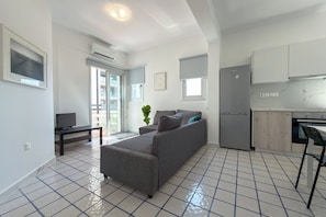 A modern living room with a grey sofa, a small table, and a TV. There is a kitchen area with cabinets and a fridge. - Harmony Beach Modern 2BR Retreat (Limassol)