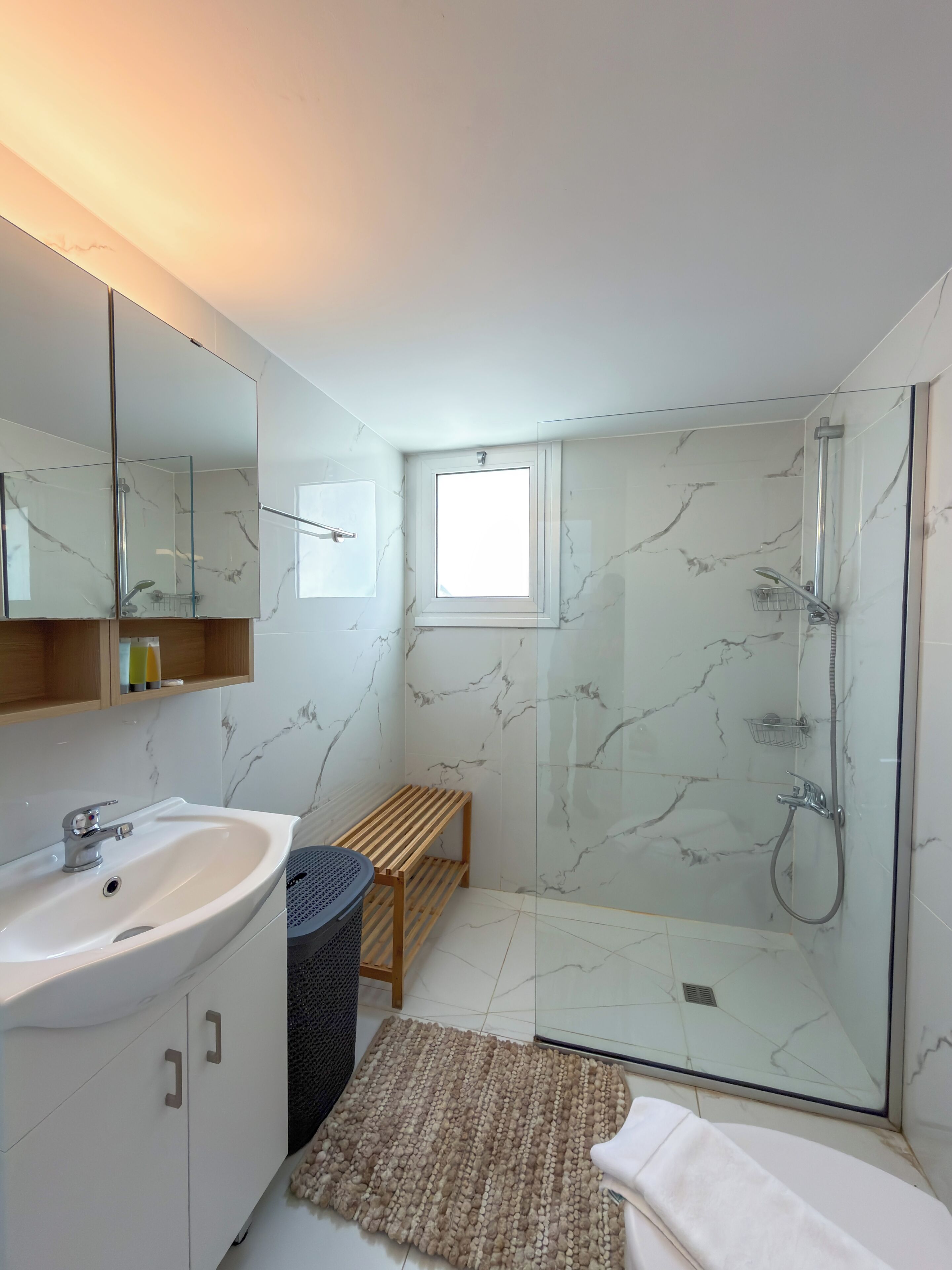 Un baño moderno con ducha envuelta en vidrio, lavamanos con encimera de mármol y un banco de madera.
