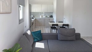 Una sala moderna con un sofá gris, cojines azul verdosos y grises, un comedor con una mesa blanca y sillas negras, y una cocina con armarios blancos y un salpicadero de azulejos.