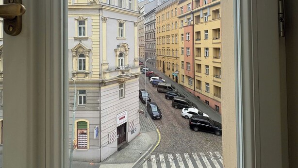 Blick durch ein Fenster auf eine Straße mit mehrstöckigen Gebäuden, abgestellten Autos und einer Fußgängerampel.