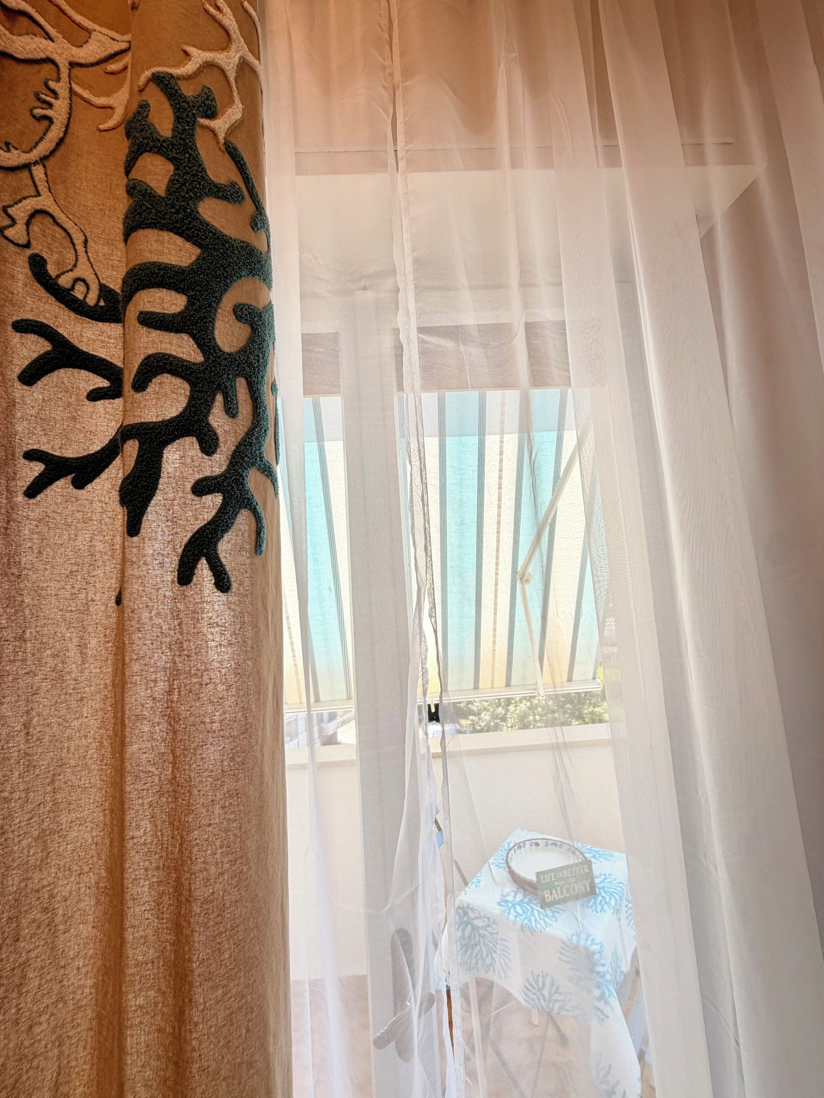 Une pièce avec un rideau blanc et léger, un rideau à motifs sur le côté gauche, et une vue sur un balcon avec une table et des chaises.