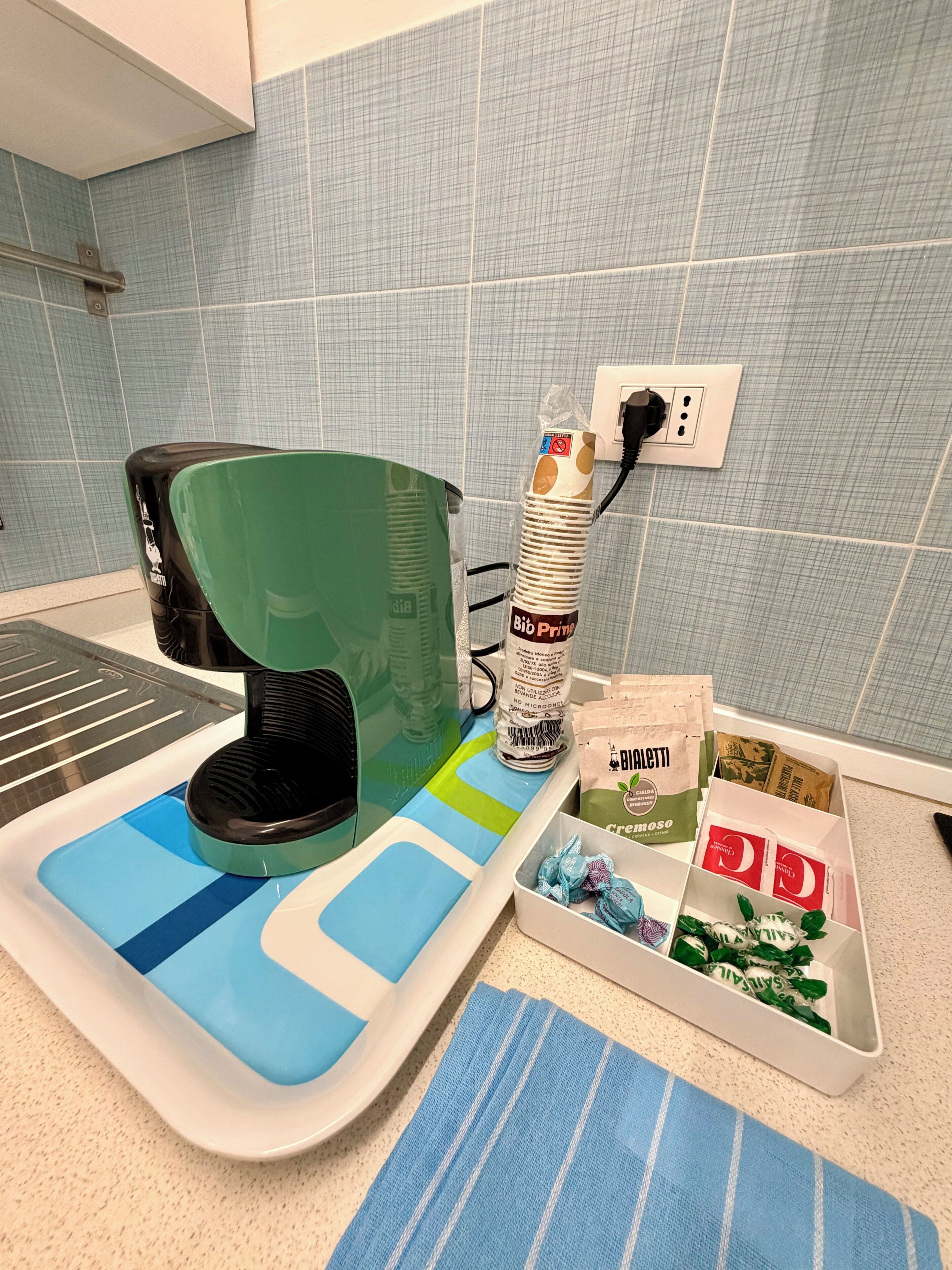 Un comptoir de cuisine avec une machine à café, une chaise verte, un tapis à rayures bleues et blanches, et un plateau contenant divers objets, notamment un paquet de café Bioaletti et un pot de crème dessert.