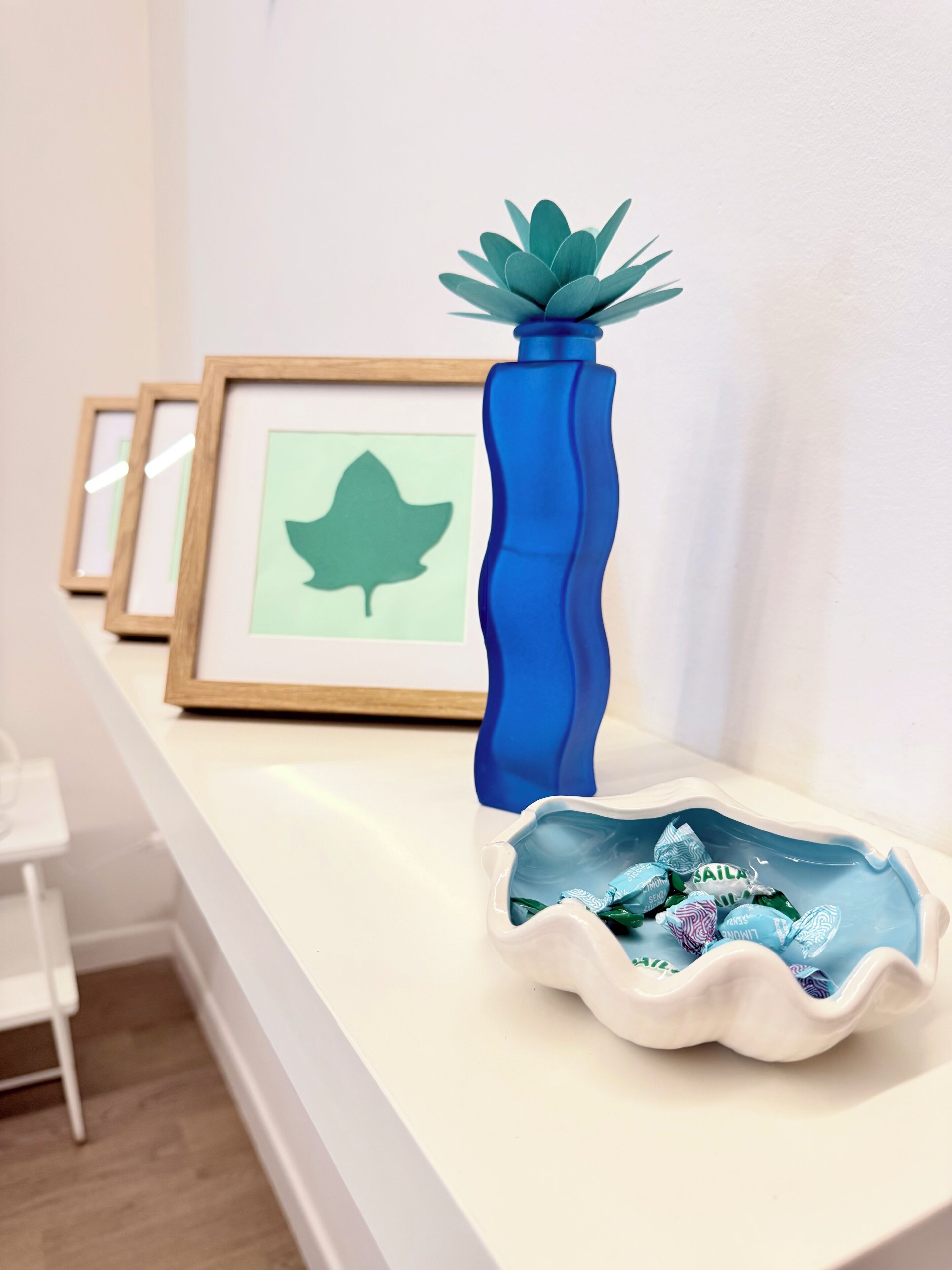 Un estante blanco con un jarrón azul y un cuenco decorativo, una obra de arte enmarcada y una silla blanca.
