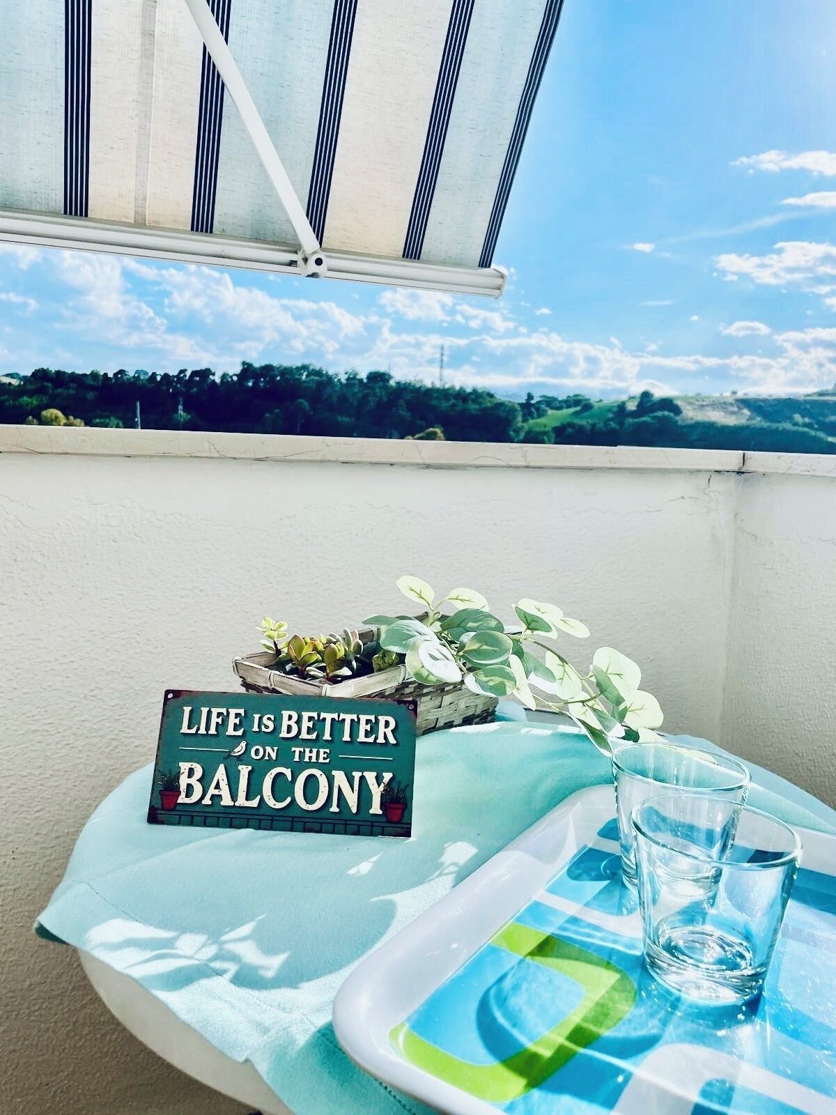 A balcony with a view of trees and a clear sky. There is a sign that reads 'Life is better on the balcony' and a glass of water on a tray.