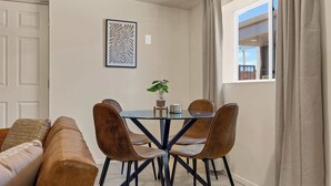 A dining area with a glass table, four chairs, a plant, and a framed artwork on the wall.