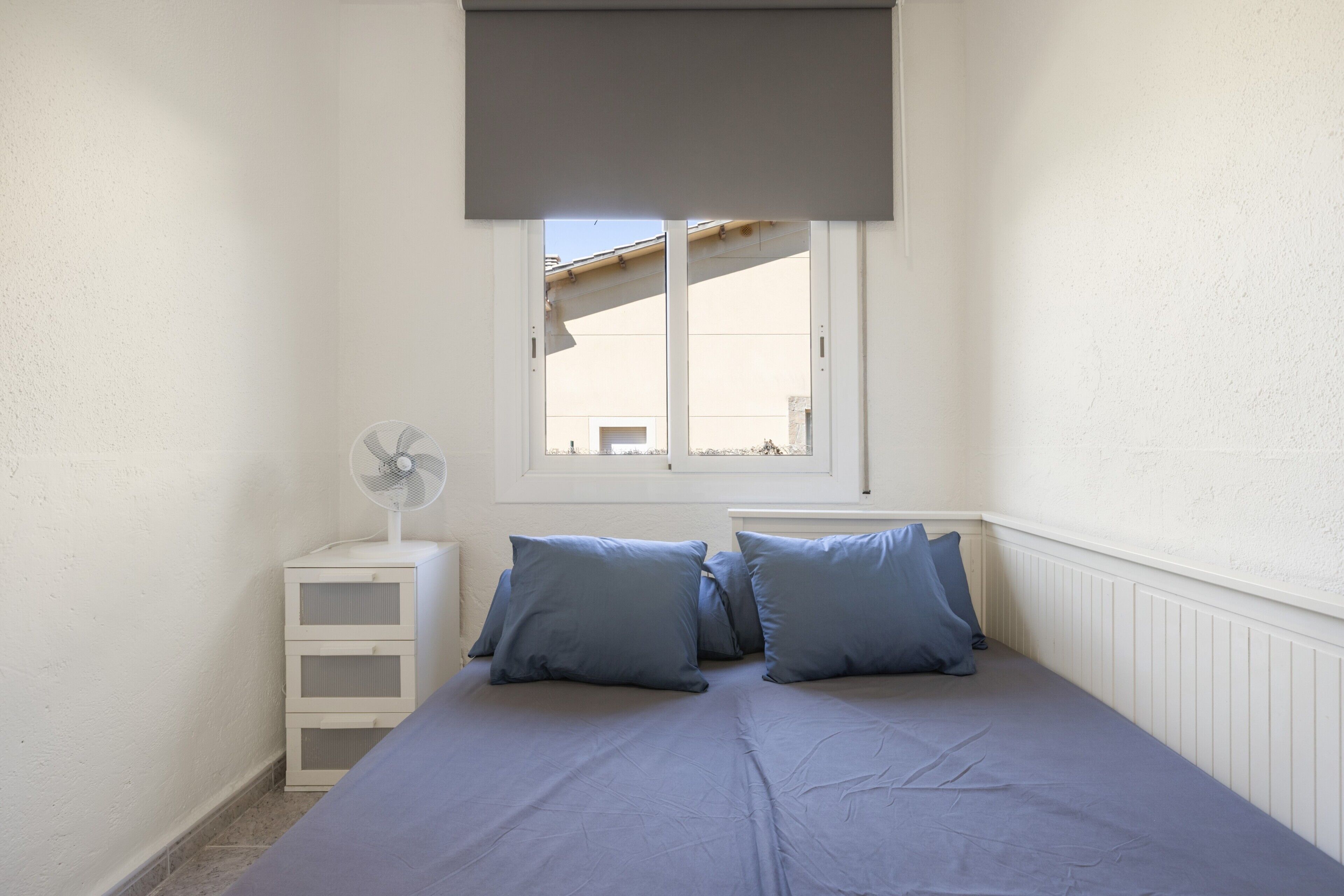 Un dormitorio con cama, dos almohadas, una mesita de noche con un ventilador y una ventana que ofrece vistas a edificios.