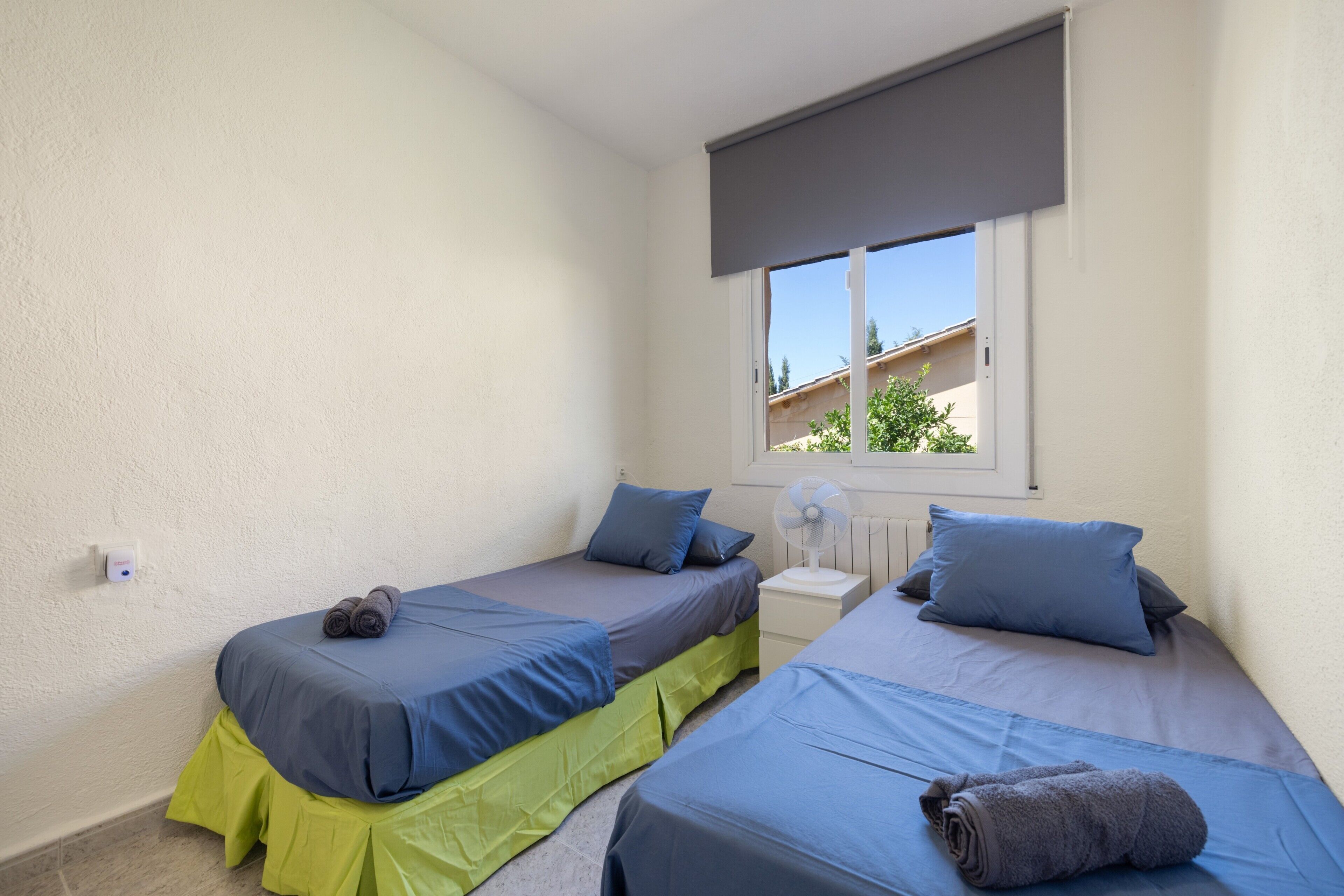 A bedroom with two beds, a window with a view, a bedside table with a fan, and a wall socket.