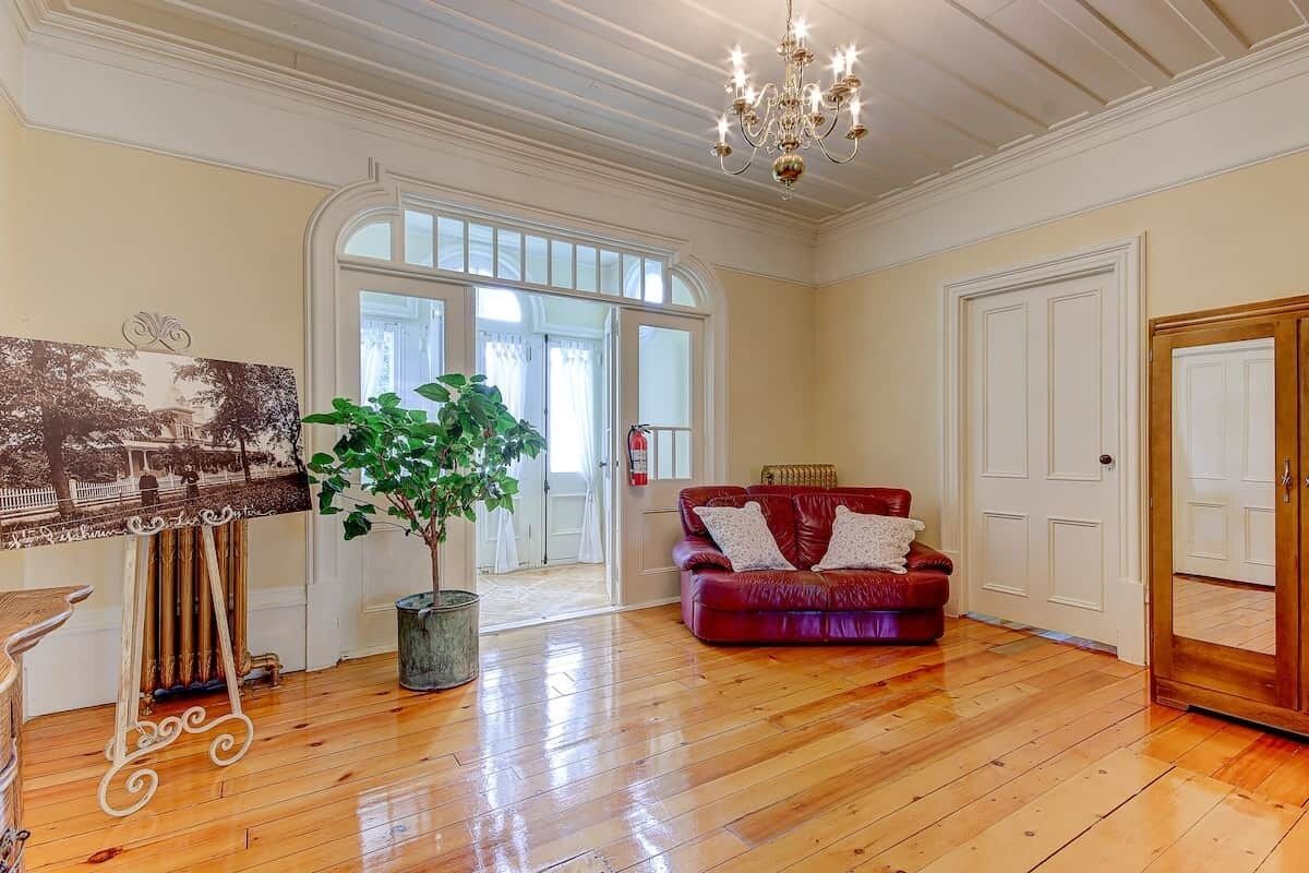 Une pièce spacieuse avec un plancher en bois, un canapé en cuir rouge, une grande plante en pot, un tableau encadré au mur et un lustre.