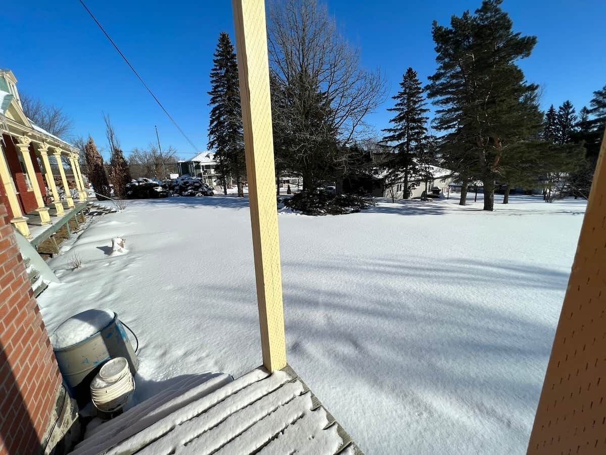 A snow-covered outdoor area with a wooden structure, possibly a porch, and a brick building in the background.