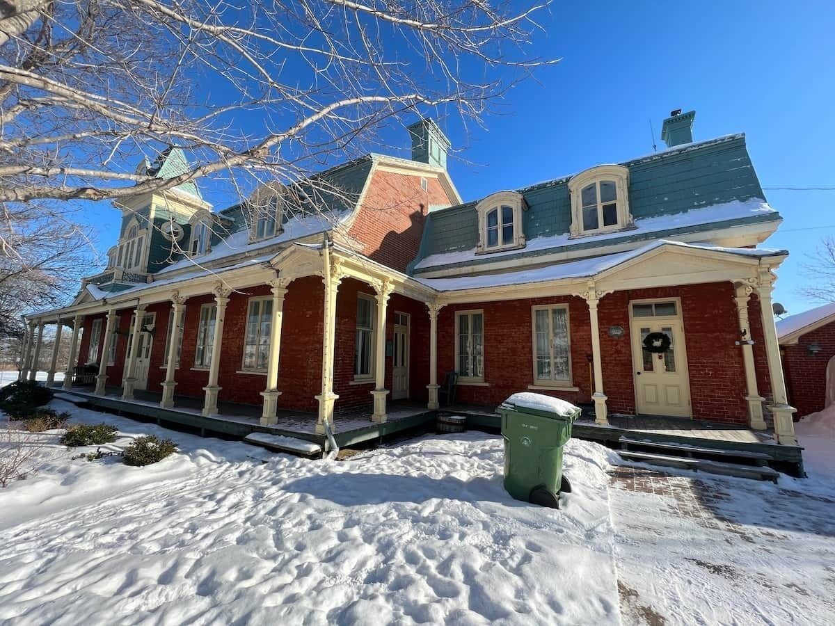 A red brick building with a green roof, a porch, and a green trash bin in front.