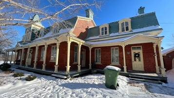 A red brick building with a green roof, a porch, and a green trash bin in front.