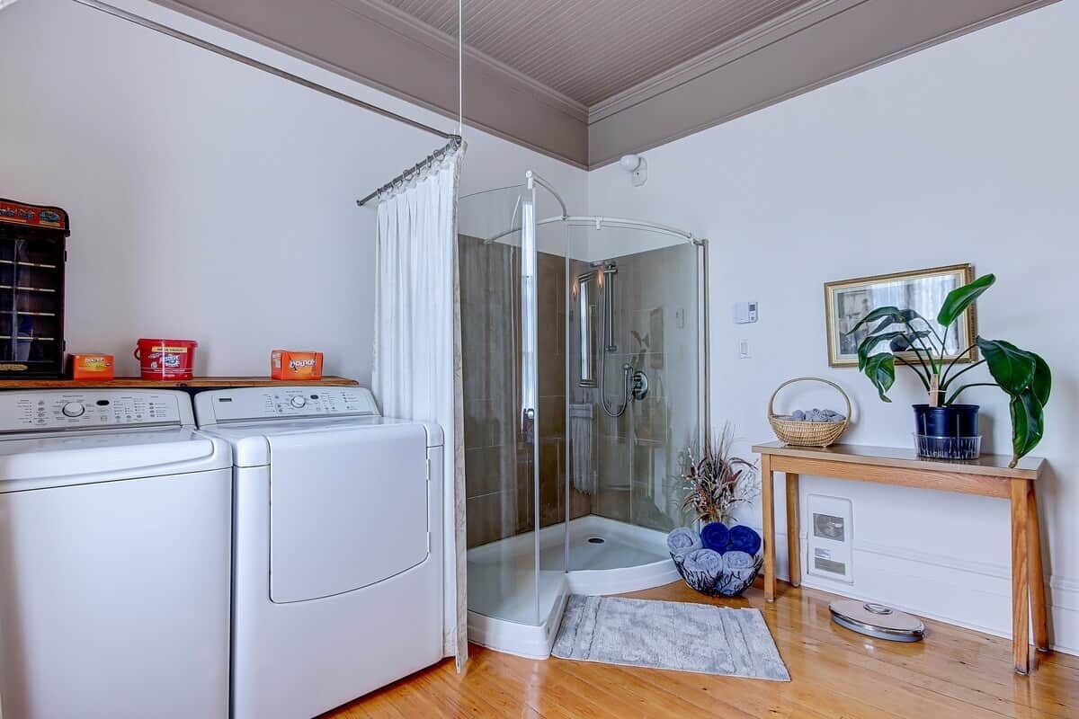 A laundry area with two washing machines, a glass shower enclosure, a wooden console table with a plant, and a framed picture on the wall.