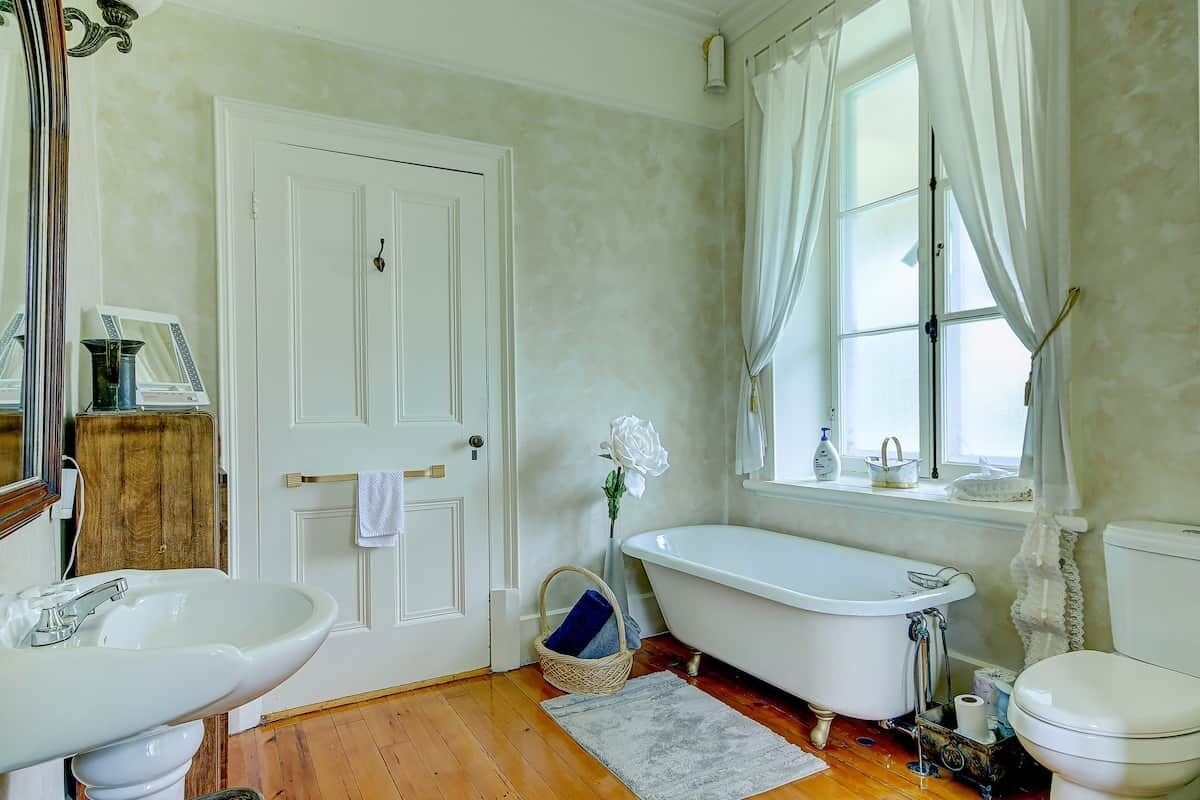A bathroom with a white door, a freestanding bathtub, a sink, and a window with curtains.