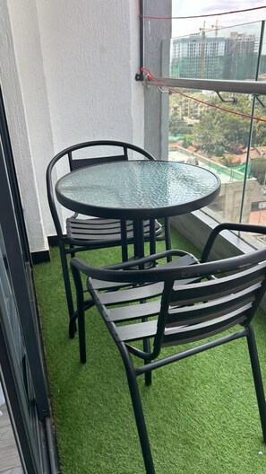 Uma pequena varanda com uma mesa de vidro redonda e duas cadeiras, rodeada por grama sintética.