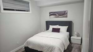 A bedroom with a bed, a nightstand, a wall-mounted artwork, and a window with blinds. - Brand new Cozy Basement Suite (Calgary)