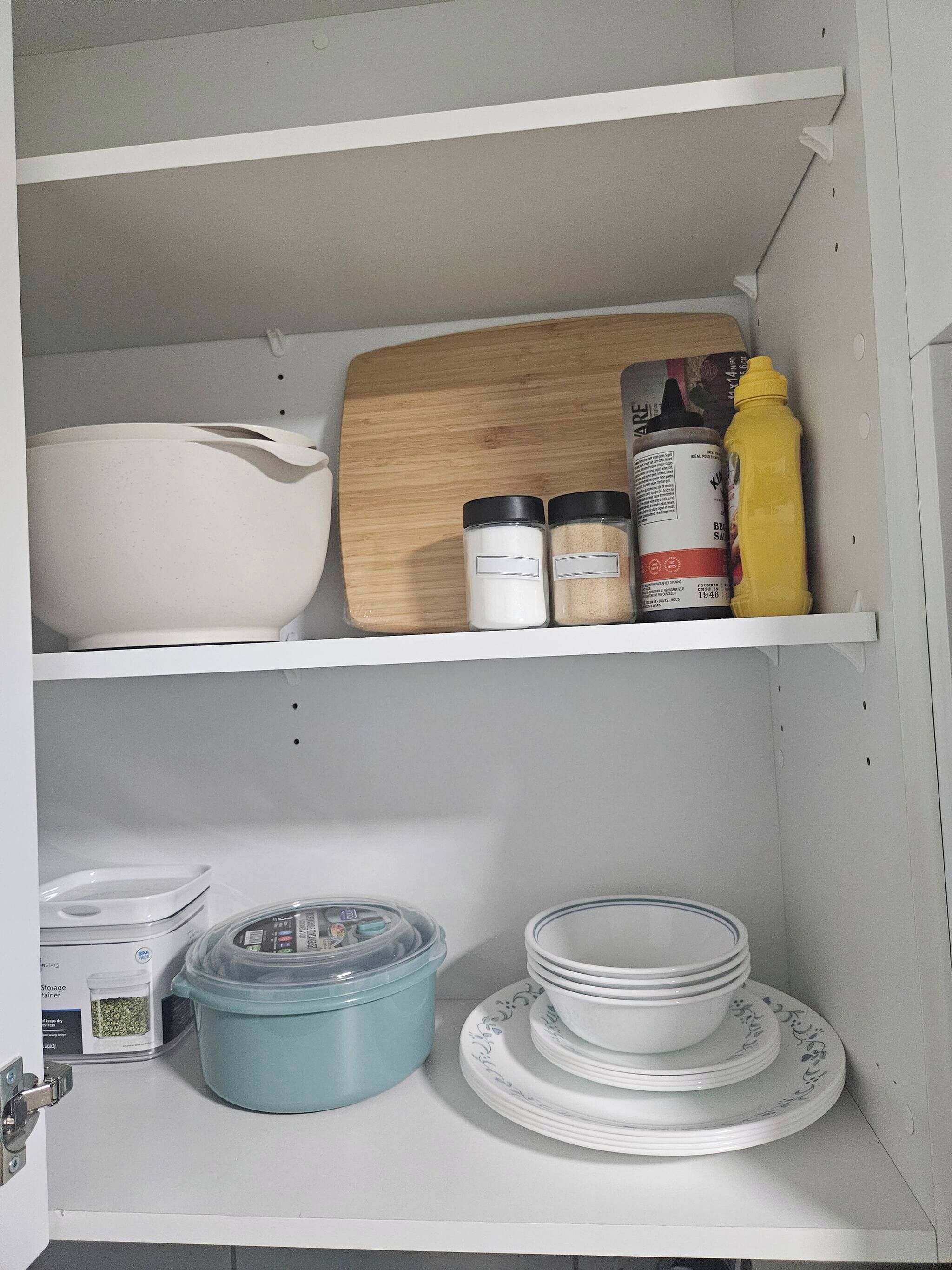Una cucina con pensili bianchi, un tagliere, barattoli, una bottiglia gialla, un contenitore blu e piatti bianchi impilati.