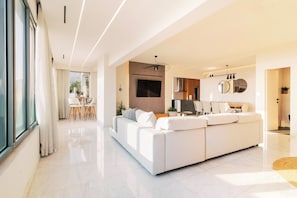 Ein modernes Wohnzimmer mit einem großen weißen Sofa, einem Essbereich mit Tisch und Stühlen sowie einer Küchenzeile mit Fernseher und Schränken.