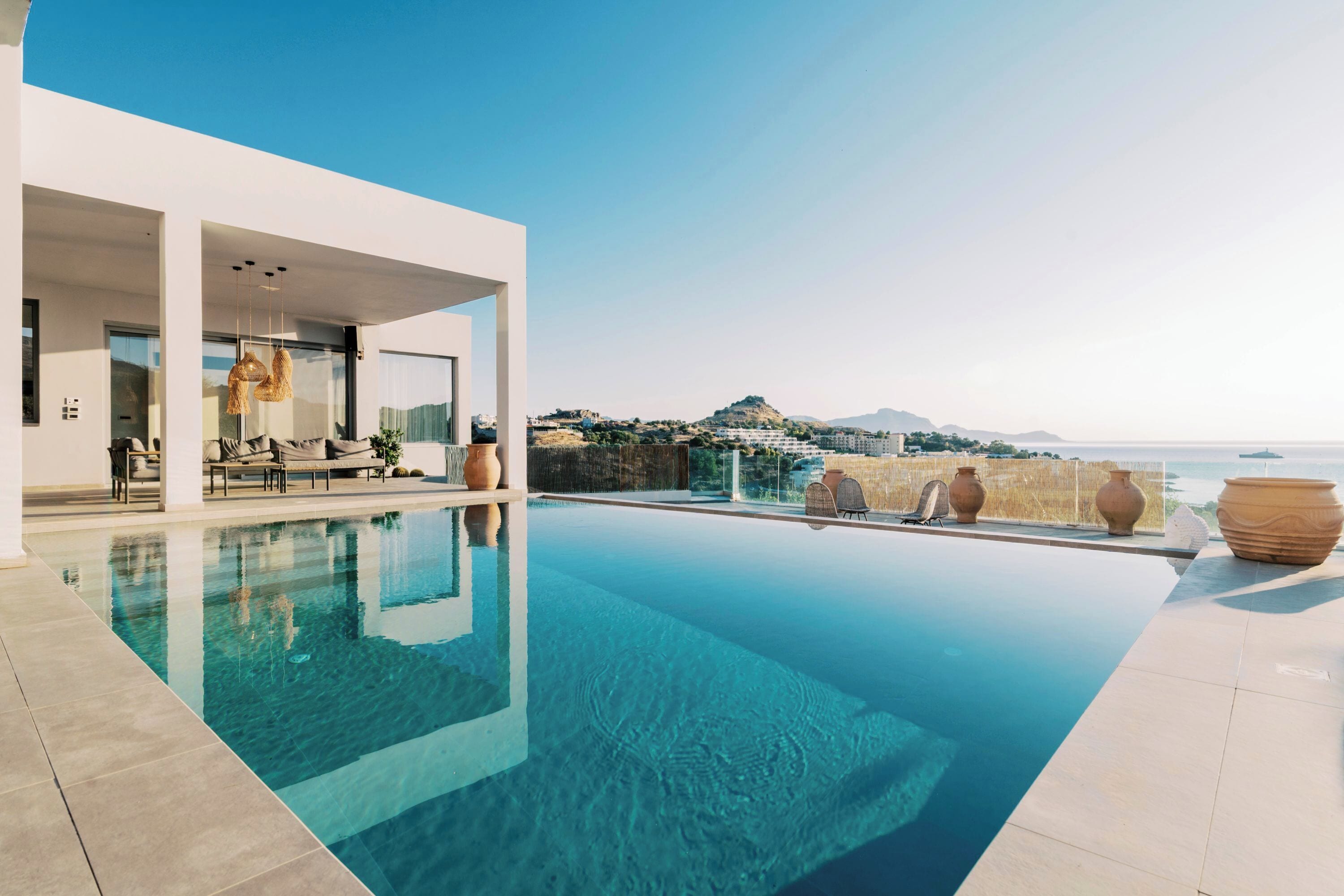 A modern pool area with clear water, a white building, and a view of the sea and hills.