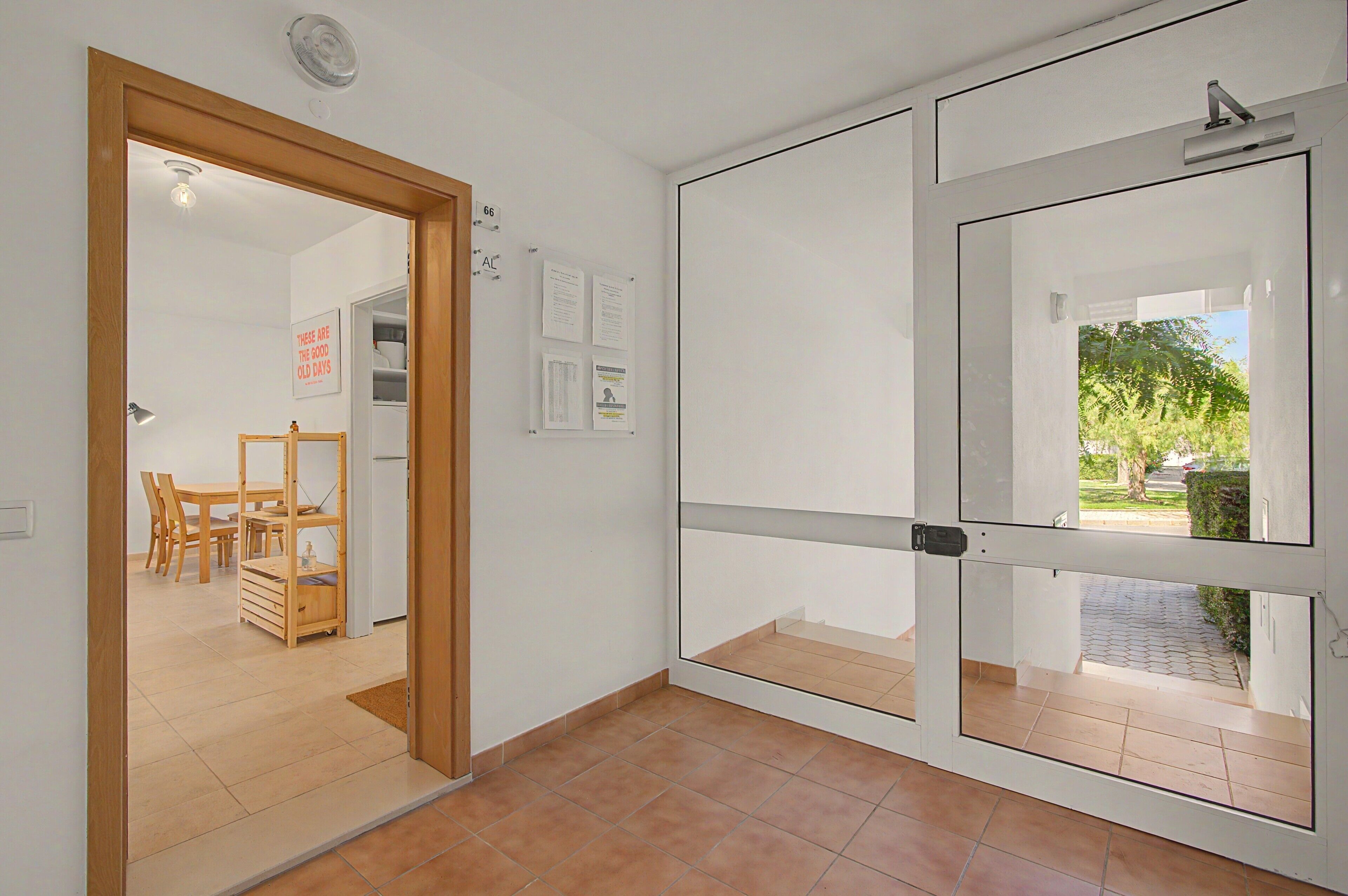 A room with a wooden door leading to a dining area, a bulletin board with notices, and a view of a garden through a glass door.