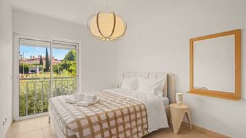 A bedroom with a bed, a bedside table, a hanging light fixture, a window, and a mirror on the wall.