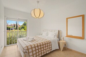 A bedroom with a bed, a bedside table, a hanging light fixture, a window, and a mirror on the wall. - Sunny 2-Bedroom Apartment in a Gated Community with Pool, 5 Minutes from the Beach (Tavira)