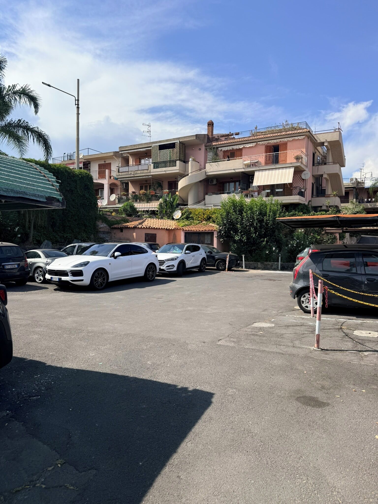 A parking lot with several parked cars, a building with balconies and a spiral staircase, and a chain-locked vehicle.
