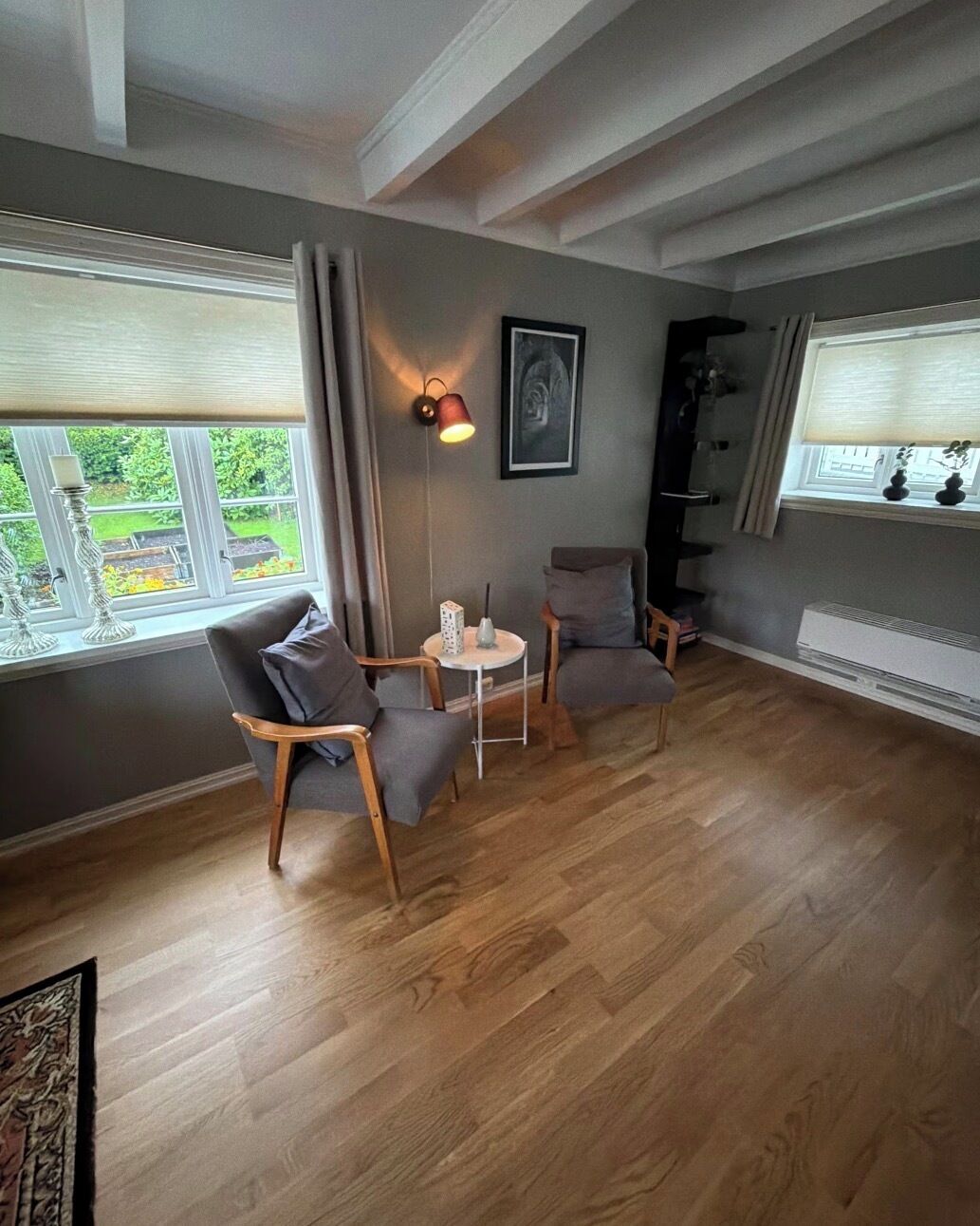 Una habitación con suelo de madera, dos sillones grises, una mesita redonda, una lámpara de pared y un cuadro enmarcado.