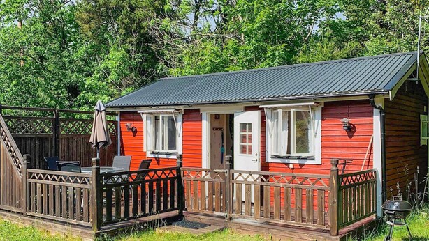 Eine kleine, rote Holzhütte mit Veranda und Grill, umgeben von viel Grün.