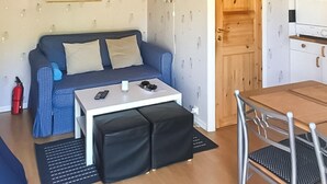 Ein kompakter Raum mit einem blauen Sofa, einem kleinen weißen Tisch, einem schwarzen Hocker, einem Metallstuhl und einer Holztür.
