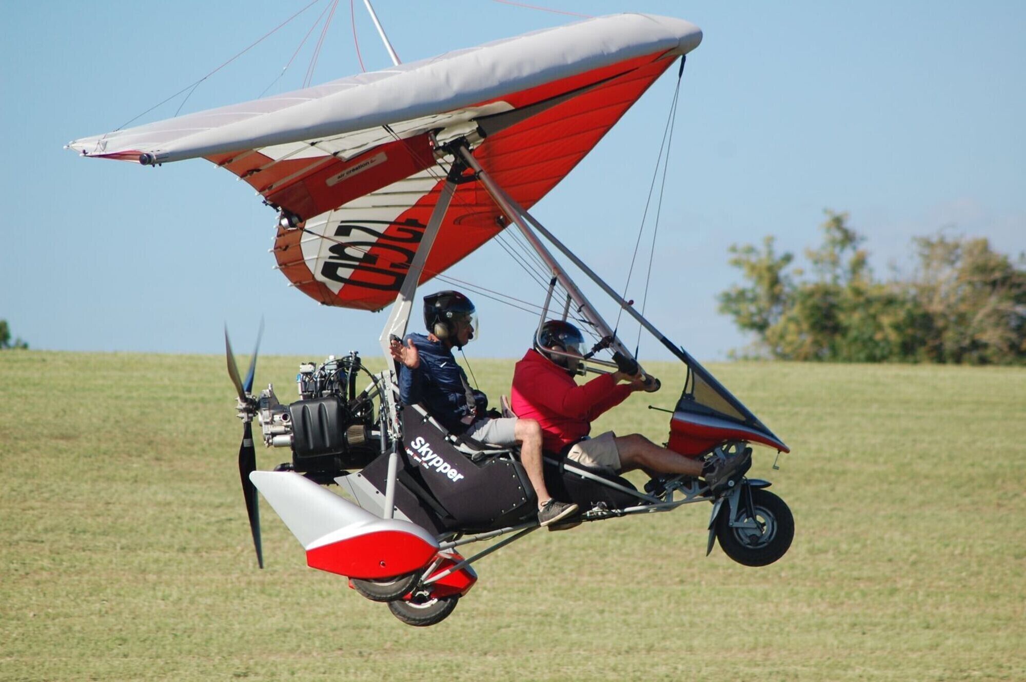 Un deltaplane avec deux passagers à bord, l’un portant un t-shirt rouge et l’autre un t-shirt bleu, survole un champ verdoyant.