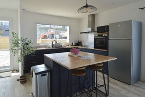 A modern kitchen with a central island, bar stools, a stainless steel refrigerator, and a window with a view of construction outside. - L'Horizon, breathtaking view, right next to a bunker (Saint-Côme-de-Fresné)