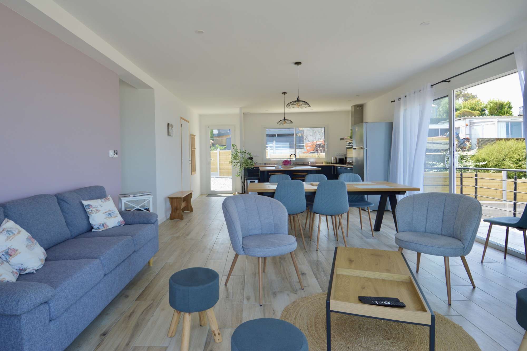 Un salon moderne avec un canapé, des fauteuils, une table basse et un coin repas, la cuisine étant visible à l’arrière-plan.