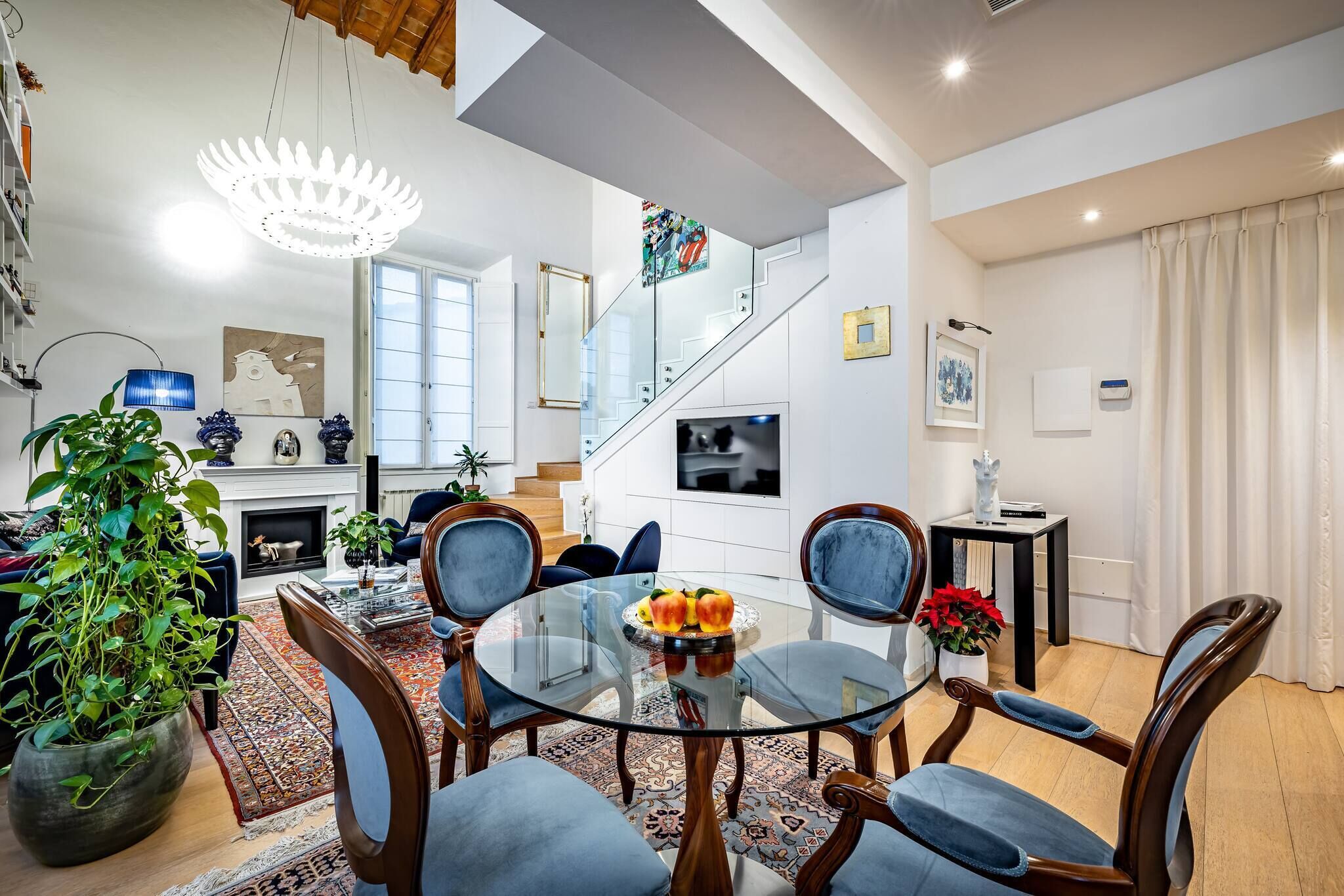 A modern living room with a glass dining table, blue chairs, a fireplace, and a staircase.